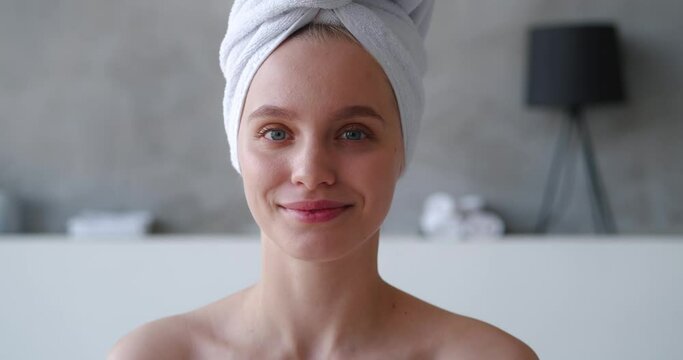 Confident close-up beauty portrait of attractive sensual blonde woman with head wrapped in white terry towel looking to the sides then smiling posing for camera in front of home bathroom background
