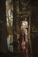 Fototapeta na wymiar Beautiful young blonde woman wearing a long red patterned skirt, standing in a water flooded forest