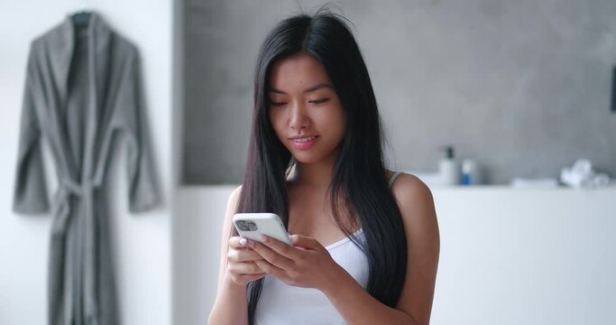 Attractive young asian woman using smartphone while standing in bathtub at home. Millennial girl in bath with mobile phone surfing social media enjoy daily bodycare in bathroom