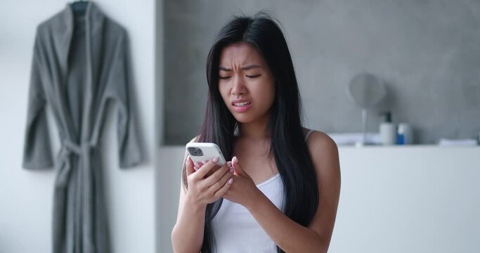 Shocked asian woman reacting on bad message on smartphone standing in bathtub. Unpleasantly surprised girl upset with a loss on mobile phone on the background of the home bathroom