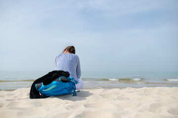 Dziewczyna, kobieta z plecakiem siedzi na piaszczystej morskiej plaży. Wpatruje się w horyzont. Lato, pastelowe kolory, przydymione błękitne niebo. - obrazy, fototapety, plakaty