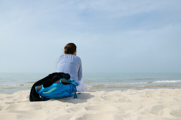 Dziewczyna, kobieta z plecakiem siedzi na piaszczystej morskiej plaży. Wpatruje się w horyzont....