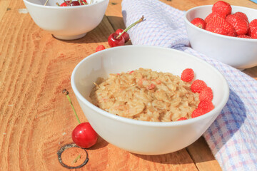 Oatmeal with wild strawberries in a plate on a wooden table. Strawberries in a white plate and a cherry next to it. Healthy breakfast or snack.