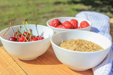 Oatmeal, cherries and strawberries in white separate plates on a wooden table. The concept of healthy eating, the guarantee of health. Summer food composition.