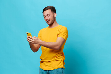 Enjoyed tanned handsome man in yellow t-shirt chatting with friends online shopping using phone posing isolated on blue studio background. Copy space Banner Mockup. Online communication concept