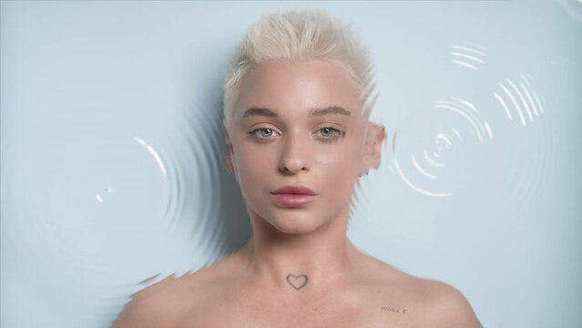 Beautiful young slim blond Caucasian woman turns to the camera looking at it through rippling water on pale blue background | Face hydration concept