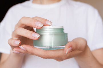 Female hands with natural pink manicure holding a transparent cream jar. Woman in white holds...