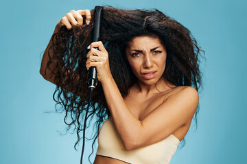 Angry confused upset curly Latin woman using a hair straightener posing isolated on blue wall...