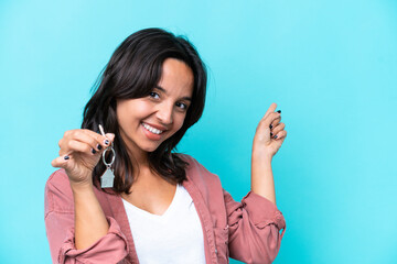 Young hispanic woman holding home keys isolated on blue background pointing back