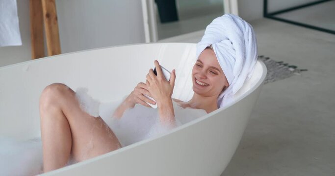 Young Charming Woman With A White Terry Towel On Head Lying Naked In Bathtub, Smiles Using Mobile Phone While Taking Bubble Bath, Enjoying Beauty Spa Day At Home Bathroom. Body And Skin Care Concept