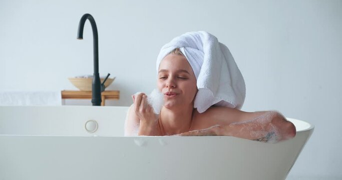 Attractive gorgeous blonde woman with closed eyes of pleasure and bath towel wrapping her hair, smiling lying in bathtub and taking bubble bath, playing with foam at home bathroom. Skin and body care