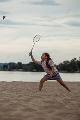 Side view of little concentrated girl with long dark hair wearing white T-shirt, blue skirt, playing badminton on sandy beach of river. Child raising racket, preparing to hit shuttlecock. Vertical.