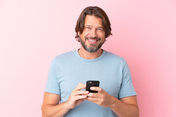 Senior dutch man isolated on pink background looking at the camera and smiling while using the...