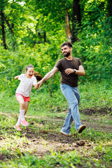 Naklejka na ściany i meble Portrait of happy family walking in park forest around green trees, having fun. Little cheerful daughter holding hand of middle-aged smiling bearded man father. Girl skipping. Love, summer activities.