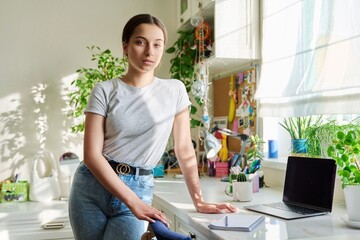 Teenage girl looking at camera, standing at home in room with laptop on table