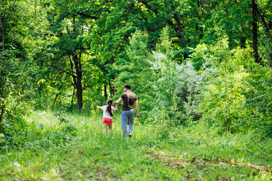 Portrait of wonderful family walking in forest around green trees. Little daughter holding hand of middle-aged bearded father. Surprised girl looking at man attentively. Summer activities, vacation.