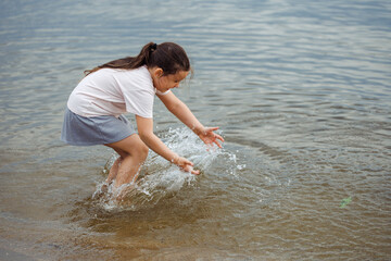 Fototapeta premium Side view of little smiling wonderful girl with long dark hair wearing white T-shirt, blue skirt, standing in water river lake, having fun. Holiday, weekend, vacation, summer activities. Vertical.