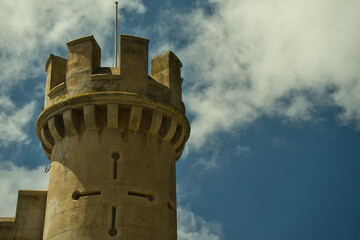 Top close-up of a castle tower against blue sky and white clouds background.