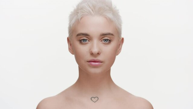 Attractive young slim blonde Caucasian woman with bare shoulders moves her head up, opens her eyes and looks at the camera on white background | Droopy eyelids removal concept