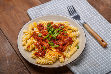 A plate with bolognese pasta on a wooden table next to a light napkin and a fork.