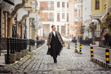 Fototapeta premium Young elegant man actor dressed in long coat and classic tailcoat on background of City. Fashion style guy with wine bottle. Street style male portrait looking to camera. Modern urban walk in town