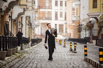 Young elegant man actor dressed in long coat and classic tailcoat on background of City. Fashion style guy with wine bottle. Street style male portrait looking to camera. Modern urban walk in town