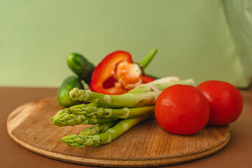 Vegetables lie on a wooden board: tomatoes, asparagus, cucumbers, red bell peppers. brown, light green background. place for text.