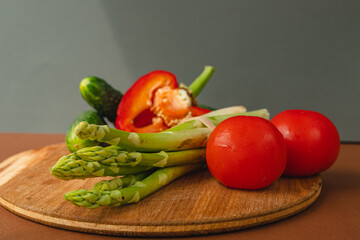 Vegetables lie on a wooden board: tomatoes, asparagus, cucumbers, red bell peppers. brown, dark gray background. place for text.