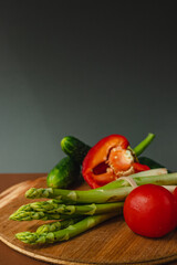 Vegetables lie on a wooden board: tomatoes, asparagus, cucumbers, red bell peppers. brown, dark gray background. place for text.