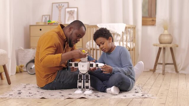 Concentrated african american father and son using screwdriver for fixing toy robot during weekend at home. Friendly family spending free time together while playing on floor in children room.