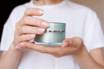 Female hands with natural pink manicure holding a transparent cream jar. Woman in white holds...