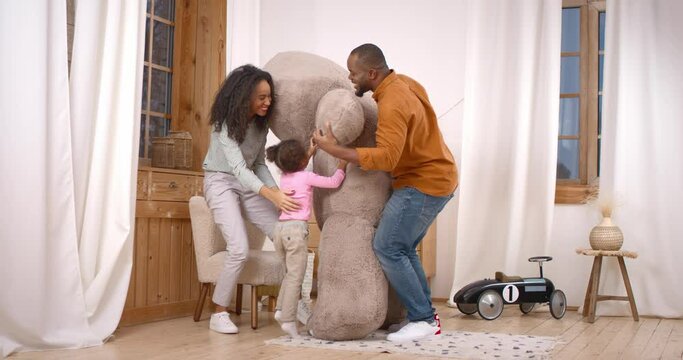 Young father unexpectedly surprising little daughter and mother with teddy bear toy in bright playroom. African american kid and loving mom cuddling dad and feeling grateful for great gift.