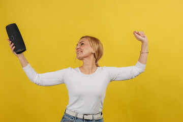 Mature woman smiling, dancing with wireless portable speaker in studio on yellow background. Music, dance concept