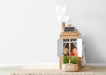 Toy cardboard house, basket with grass and balloons near light wall