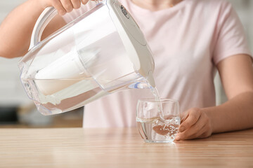 Woman pouring purified water into glass from filter jug at table in kitchen, closeup