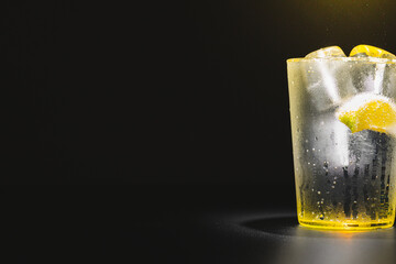 Large glass of gin and tonic, sparkling water, tonic soda with ice and a slice of lemon with a dark background and vertical lighting.
