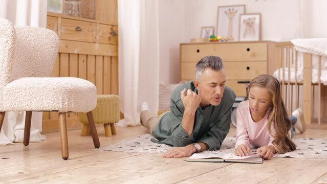 Affectionate father spending time with cute daughter for reading book at home. Handsome caucasian man and pretty preteen child lying together on floor with interesting literature in hands.