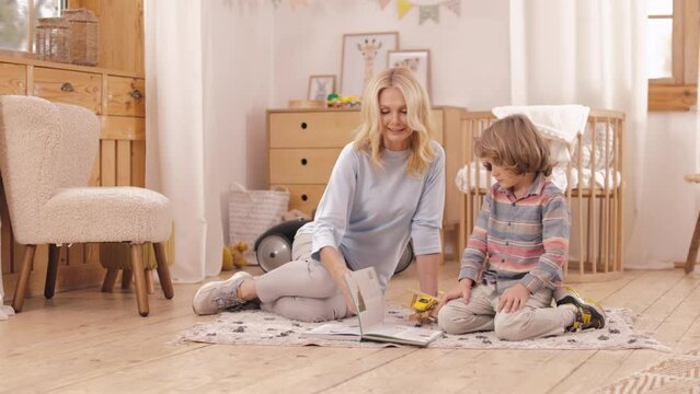 Cute little boy and his charming mom spending time at home for reading interesting book together. Caring mother showing images to preschooler son while sitting together on floor.