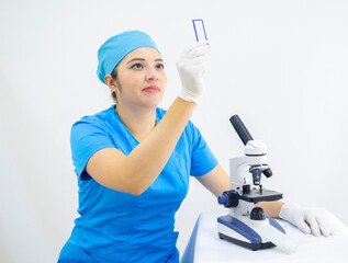 beautiful female lab technician wearing uniform and blue surgical cap, latex gloves, analyzing samples with the microscope, on white background