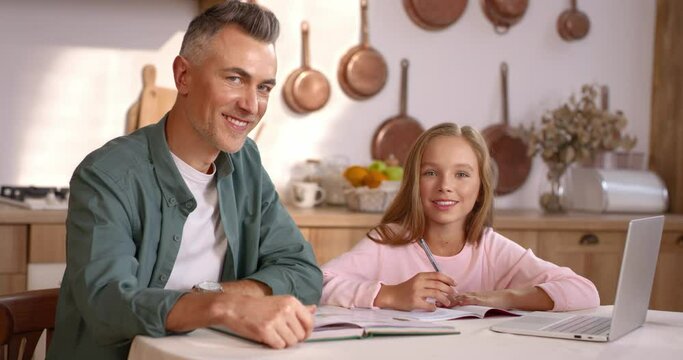 Portrait of loving caucasian dad and pretty daughter looking on each other while doing homework at kitchen. Adult male parent and kid girl smiling on camera at home.