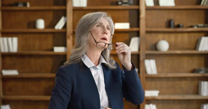 Pensive mature woman in formal wear taking off glasses and looking away with calm face. Female librarian getting lost in thoughts while standing at work with bookshelves on background.