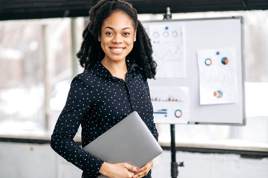 Portrait of successful young adult pretty, confident african american woman, financial top manager, wearing business shirt, stands in a modern office near markerboard, looks at camera, smiles friendly
