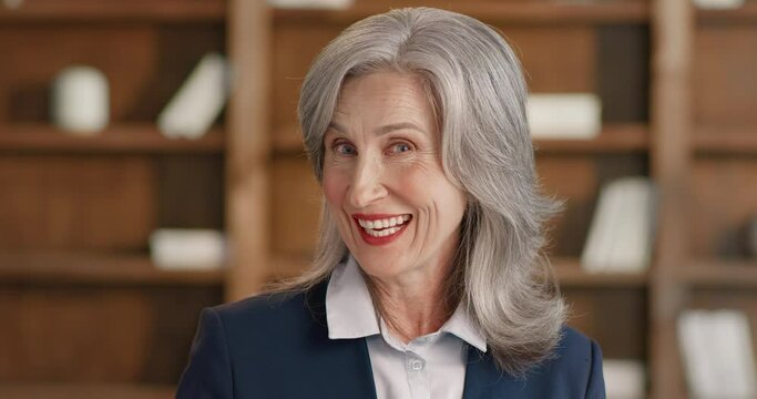 Portrait of confident mature woman talking on camera while standing at library with bookcase on blur background. Excited grey haired lady sharing impression about new book.