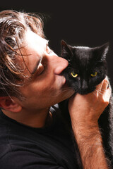  young man kissing a black cat with yellow eyes, wearing black t-shirt on black background.