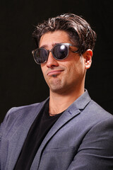 Obraz premium smiling young man looking at the camera with black sunglasses, wearing a black T-shirt and a blue jacket, on black background.
