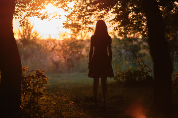 Back view of young woman in summer dress walking alone through evening dark forest
