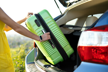 Fototapeta premium Close up of woman hands taking green suitcase from car trunk. Travel and vacations concept