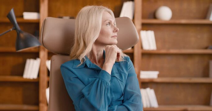Dreamy caucasian woman with blond hair sitting in armchair turning head and looking at camera. Female psychologist waiting for patient at modern room with wooden shelves on background.