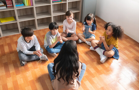 Happy Asian Female Teacher and Group Of Multiethnic Elementary School Pupils sitting on floor singing together in Classroom, Indoor activity.