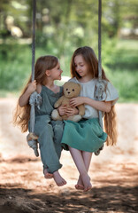 Happy laughing kids girls sisters with long hair enjoying a swing ride with a teddy bear toy on a sunny summer day